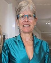 Dr. Susan Eliason smiling with short gray hair and wearing teal blue rim glasses and a teal blue wrap dress with collar