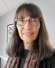 headshot photo of Dr. Erin O'Connor with long brown/grey hair and bangs smiling wearing brown rim glasses, a black top and purplish grey print scarf
