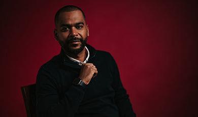 portrait photo of Harold Tavares sitting in a chair in front of a red backdrop